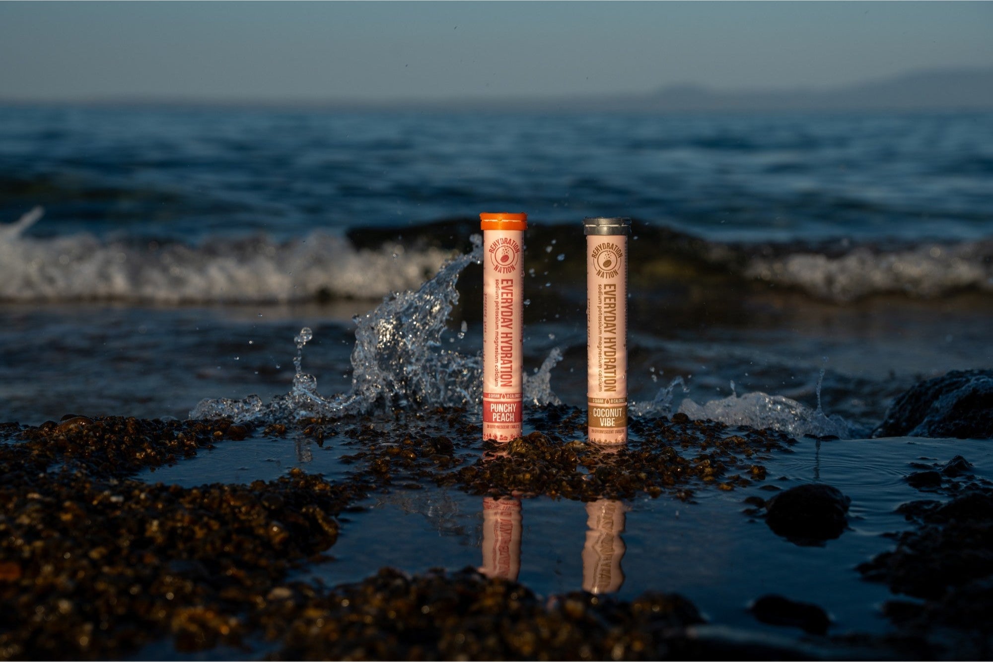 Rehydration Nation Punchy Peach and Coconut Vibe hydration tablets – tubes photographed on wet pebbles with sea waves splashing in the background